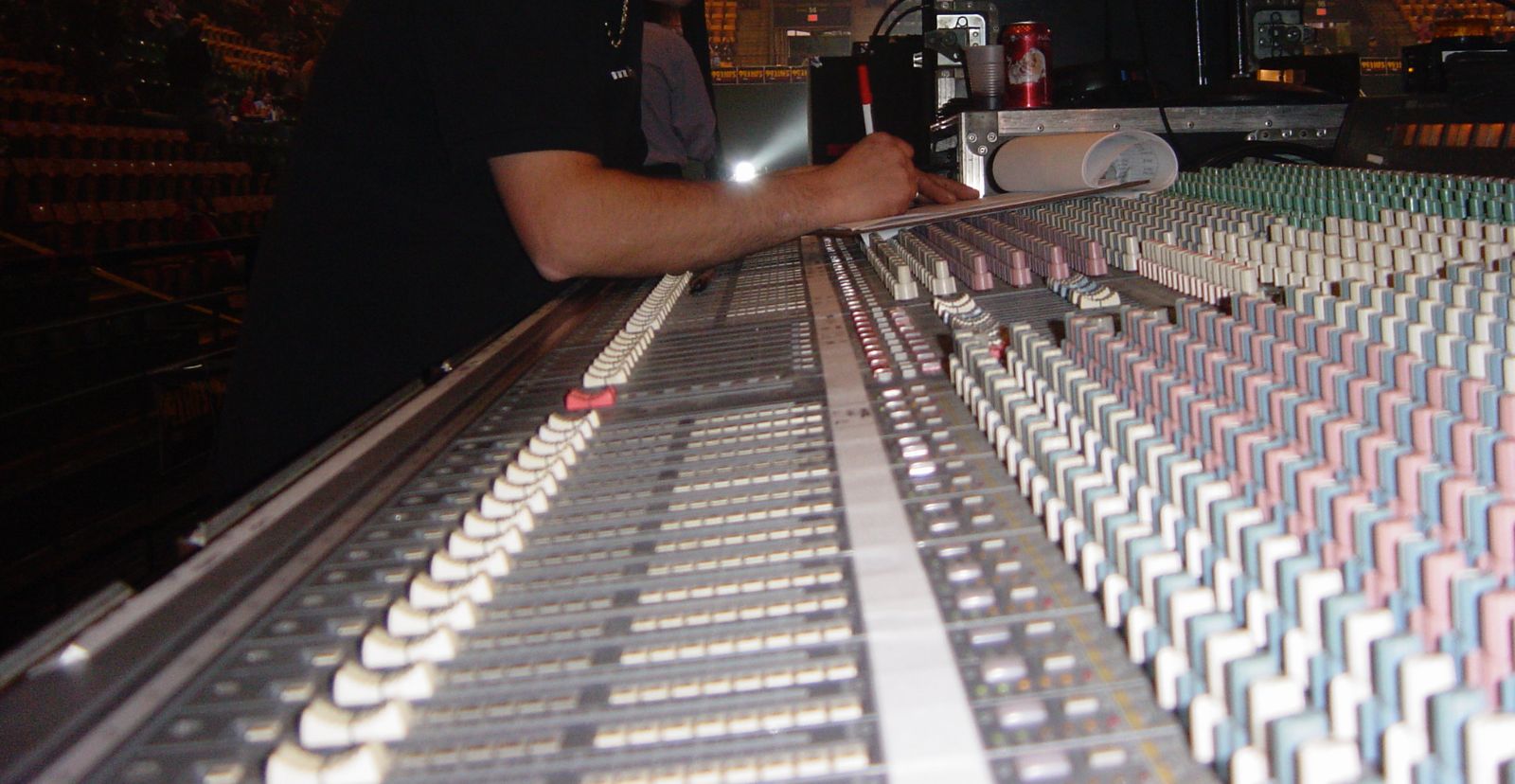 An audio tech's hands on a large analog mixing console, rows of faders and colored channel knobs stretching across the frame, with arena seats out of focus behind