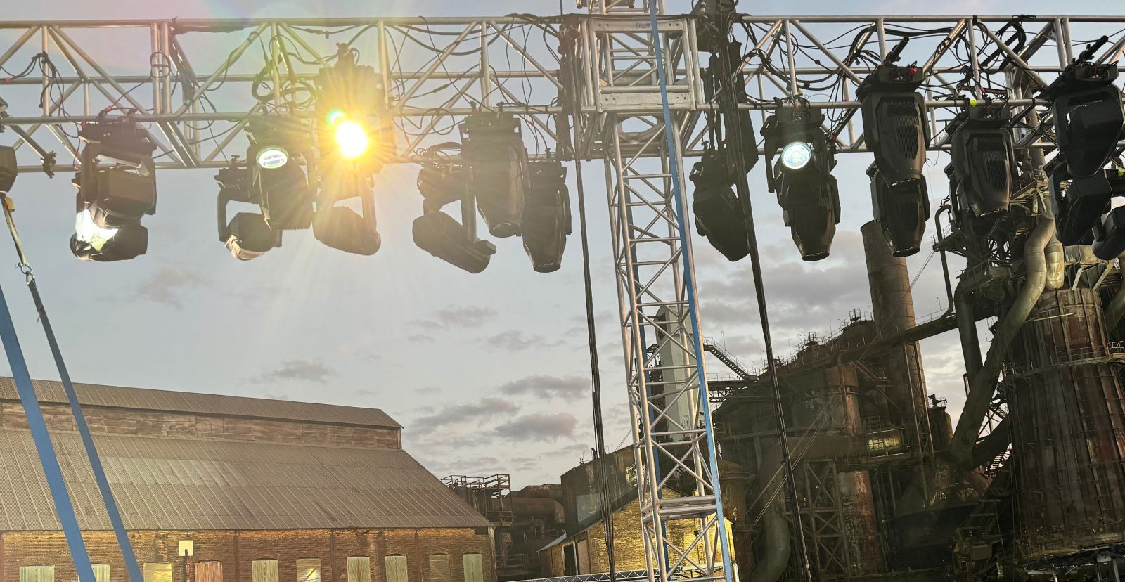 Moving-head lighting fixtures hung along an aluminum truss against a dusk sky, one fixture lit, with an old steel mill and a warehouse roof behind