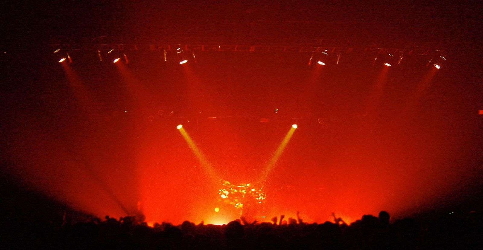 Two followspot beams from the back of a concert hall converging on a drum kit at center stage, the whole room washed in red light with the crowd in silhouette