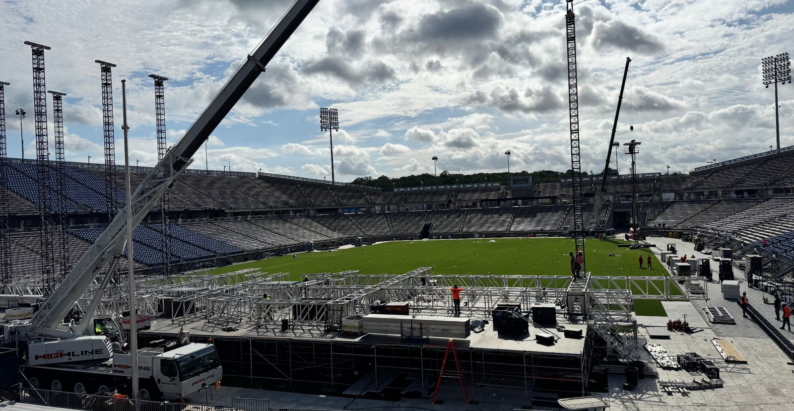 Stage build in an empty stadium: two cranes lifting sections of aluminum truss over the deck while crew in orange vests work on the structure