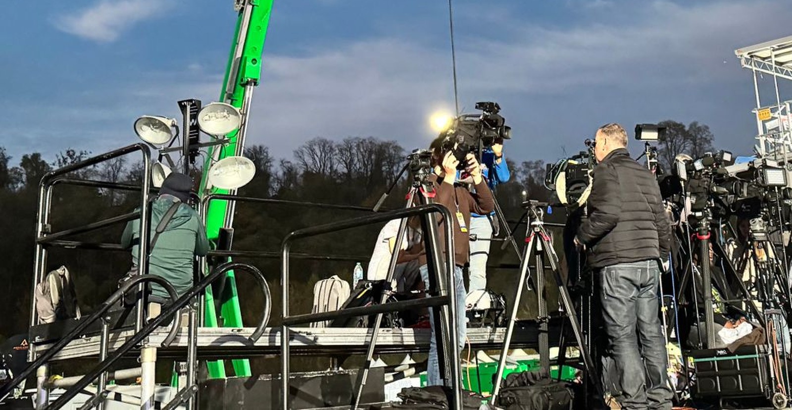 Camera operators working broadcast cameras on tripods from a raised platform at dusk, a green boom lift behind them