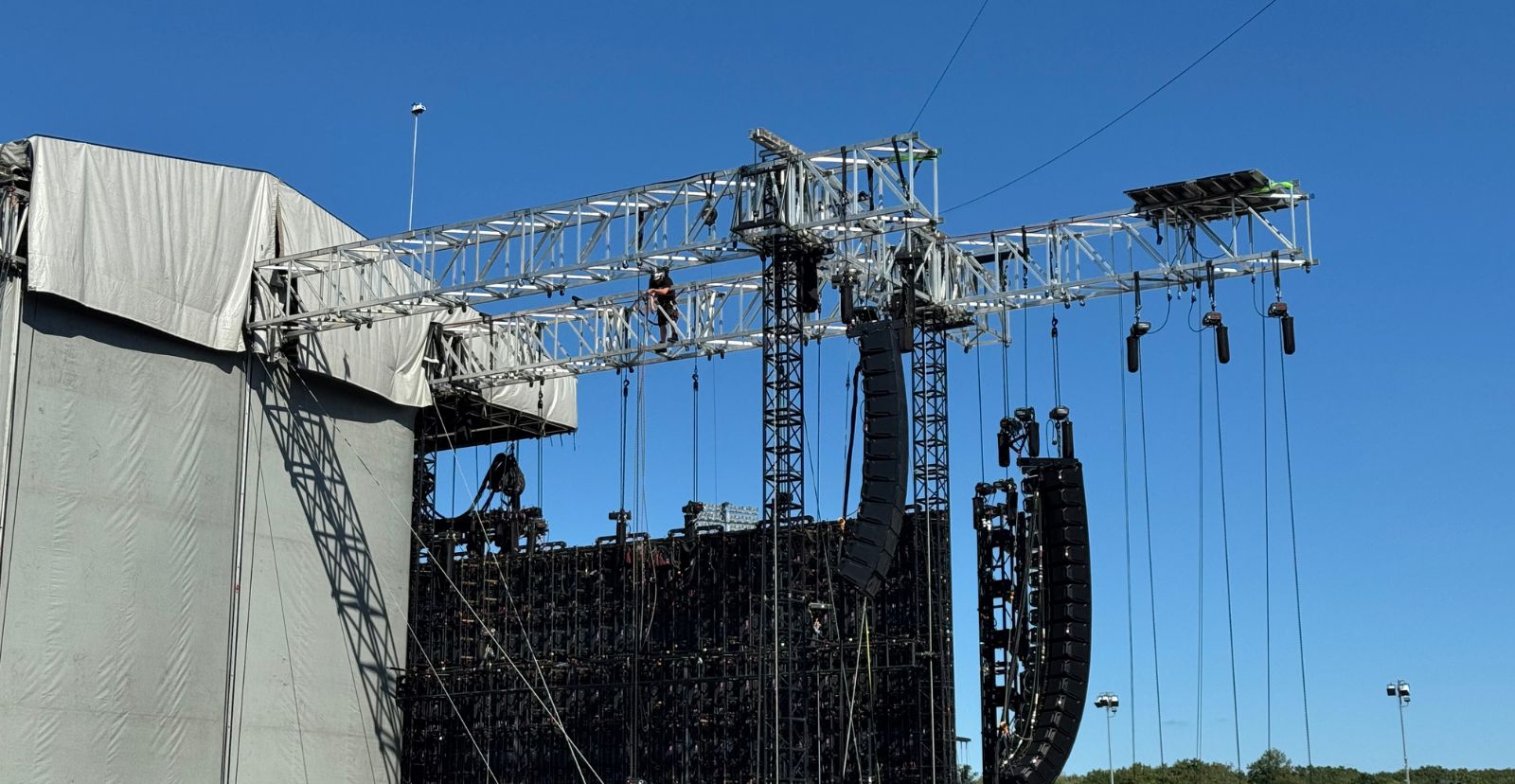 Outdoor stage roof truss with a black LED video wall and a line-array speaker hang against a clear blue sky